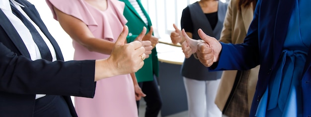 Unrecognizable unidentified female officer staff in business suit show thumb up while others standing ovation side by side applauding hands showing warm greeting celebration in blurred background.