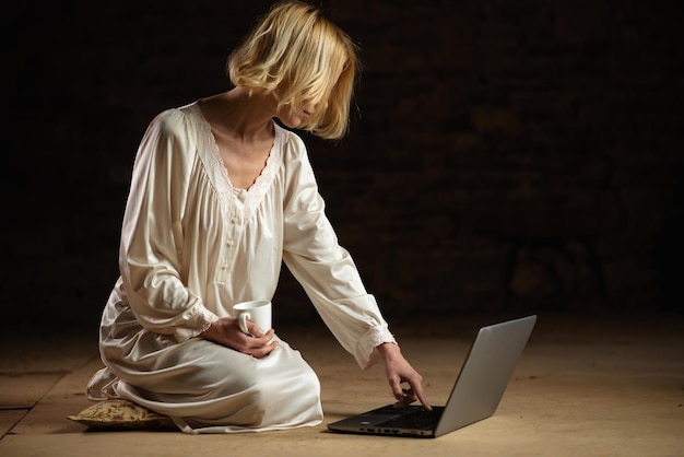 Unrecognizable woman in an empty space with coffee in her hands and a laptop on a dark background Deadline recycling concept A middleaged woman in a white nightgown