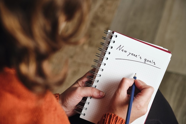 Unrecognizable woman from behind in orange sweater holds a pencil and a notebook with the words new year's goals written on it