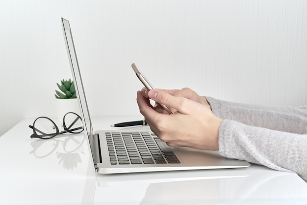 Unrecognizable woman using smartphone while typing on laptop, concept of password entry