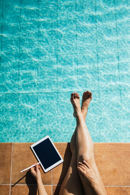 세로 형식으로 청록색 수영장 가장자리에서 태블릿을 사용하는 인식할 수 없는 여성