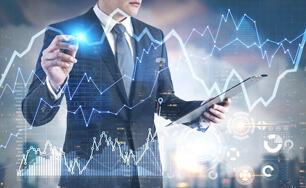 Unrecognizable young businessman is looking at his clipboard and holding a glowing marker. A blurred cityscape background, graphs and HUD. Toned image double exposure