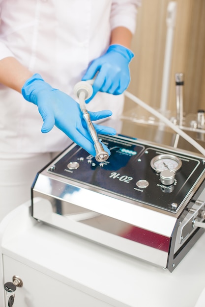 Unrecognizable young doctor with medical equipment. Female cosmetologist with dermatological apparatus of microdermabrasion in latex gloves. Polishing tool background.