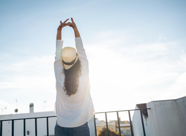 Unrecognizable young woman in hat stretching her arms to the terrace in the morning