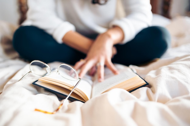 Unrecognizable young woman reading a book sitting on the bed with her glasses removed