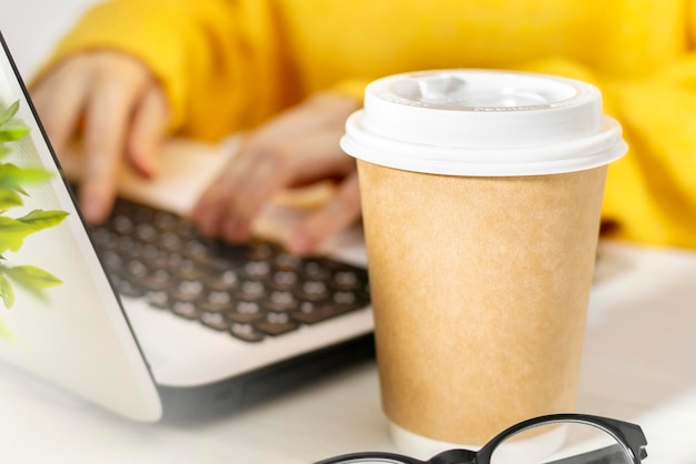 Photo unrecognizable young woman in yellow sweater working using laptop in cafe at table with cardboard coffee mug and glasses.