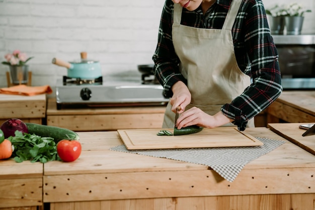 unrecognized asian woman preparing salad with vegetables on table. Healthy eating lifestyle concept. closeup lady in apron holding sharp knife cutting and chopping fresh cucumber in slice in kitchen