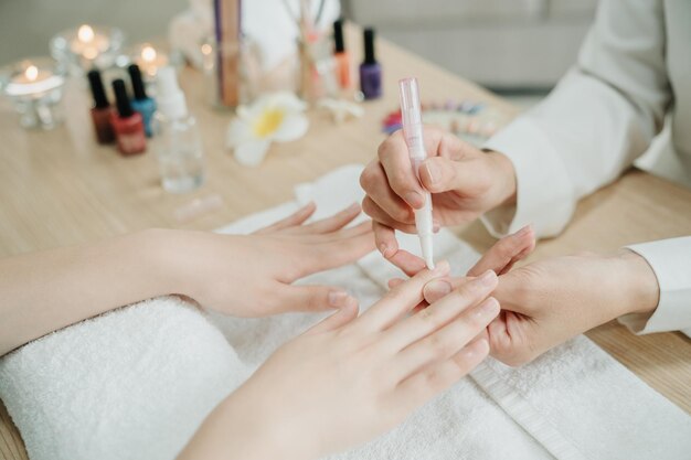 Photo unrecognized woman worker in home service holding female client hands applying nourishing oil on cuticles. beautician doing manicure work for lady customer on wooden table. close up vie concept.