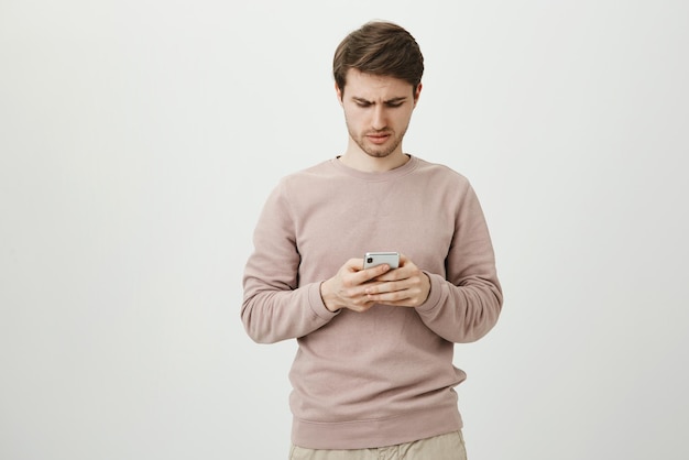 Unsatisfied gloomy good-looking european student looking at smartphone while browsing or texting and being disappointed