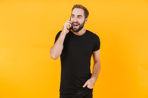 unshaved man wearing basic black t-shirt rejoicing and talking on smartphone isolated over yellow wall