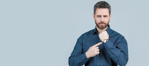 Unshaven businessman in casual shirt point finger at wrist watch grey background business hour Man face portrait banner with copy space