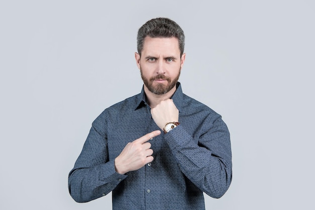カジュアルなシャツを着た無精ひげを生やしたビジネスマンは、腕時計の灰色の背景、営業時間で人差し指を指しています。