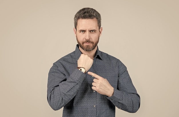 Unshaven businessman in casual shirt point finger at wrist watch grey background business hour