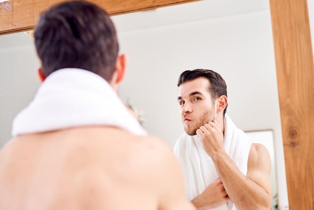 Unshaven male with white towel on his neck standing near mirror