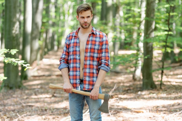 Uomo con la barba lunga in stile boscaiolo in piedi con sfondo foresta ascia