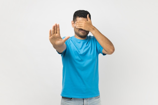 Unshaven man in t shirt standing covering his face with hand hiding his face making stop gesture