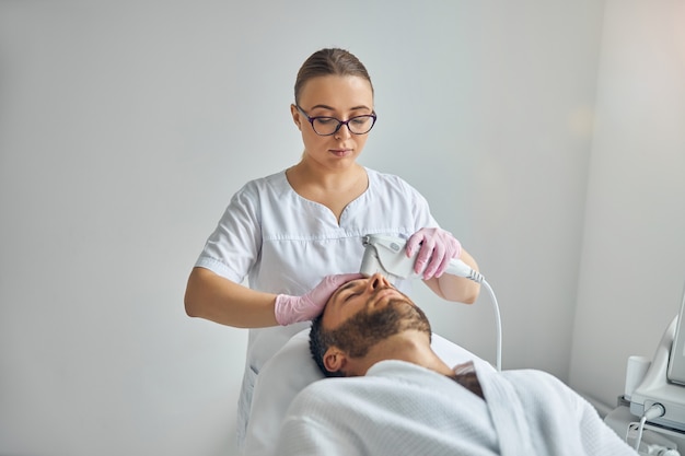 Unshaven young man lying on daybed while receiving laser facial treatment in cosmetological clinic