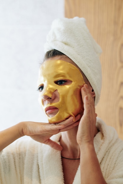 Unsmiling woman in bathrobe with golden gel sheet mask on her face looking at herself in mirror, skincare and anti-aging treatment concept