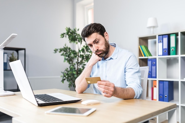 Unsuccessful online purchase Unsuccessful deal Upset young man office worker holding a credit card holding his head in shock Sitting at a desk at a laptop