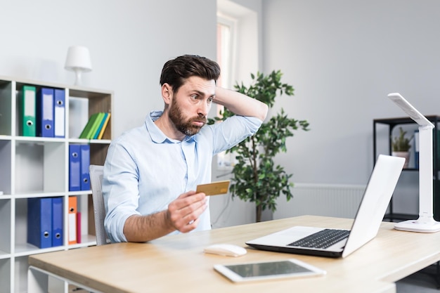 Unsuccessful online purchase Unsuccessful deal Upset young man office worker holding a credit card holding his head in shock Sitting at a desk at a laptop