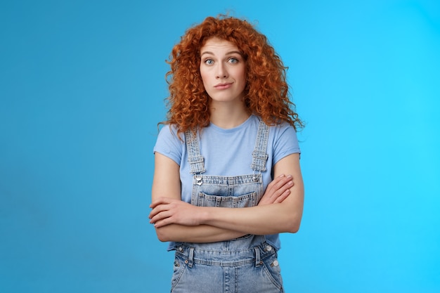 Insicuro confuso donna riccia rossa sguardo perplesso incerto braccia incrociate petto sogghignando sguardo macchina fotografica piena incredulità sospettoso dubbioso qualcuno che dice la verità in piedi salopette di jeans sfondo blu.