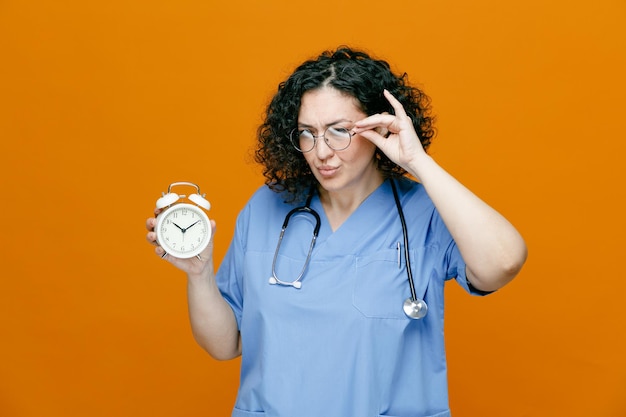 Unsure middleaged female doctor wearing uniform glasses and stethoscope around her neck showing alarm clock grabbing glasses looking at camera isolated on orange background