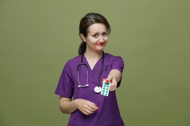 Unsure middleaged female doctor wearing uniform and stethoscope around neck stretching pack of tablets out towards camera looking at camera isolated on olive green background