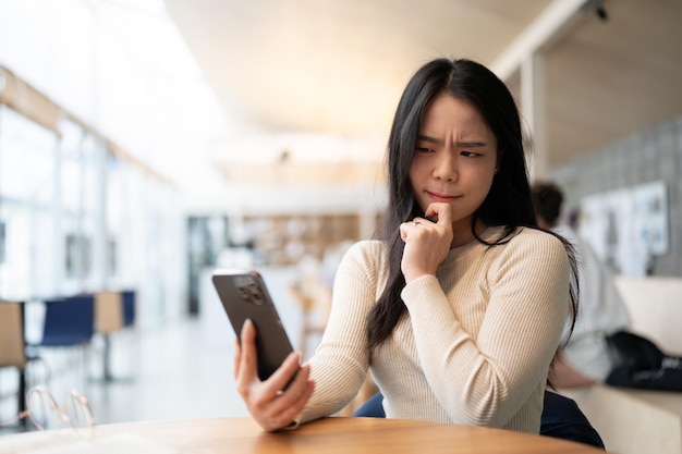 An unsure and thoughtful Asian woman is looking at her smartphone screen with a serious