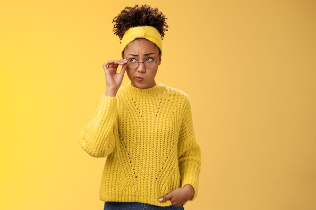 Unsure thoughtful hesitant cute african-american millennial teenage girl in round glasses sweater headband solving riddle mind look up doubtful touch eyewear smirking, thinking yellow background.