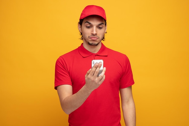 unsure young delivery man wearing uniform and cap holding and looking at mobile phone isolated on yellow background