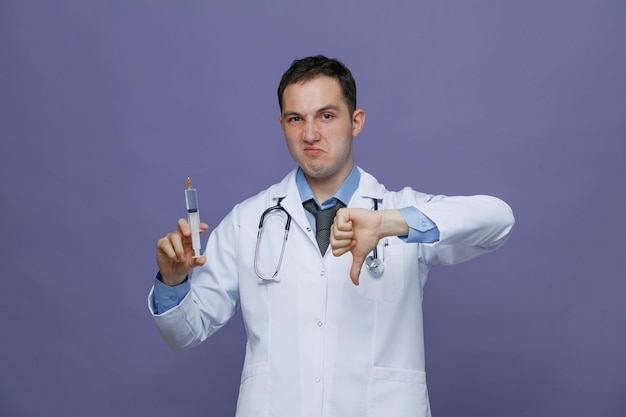 Unsure young male doctor wearing medical robe and stethoscope around neck holding syringe with needle looking at camera showing thumb down isolated on purple background