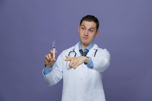 Unsure young male doctor wearing medical robe and stethoscope around neck holding syringe with needle looking at side pointing at side isolated on purple background