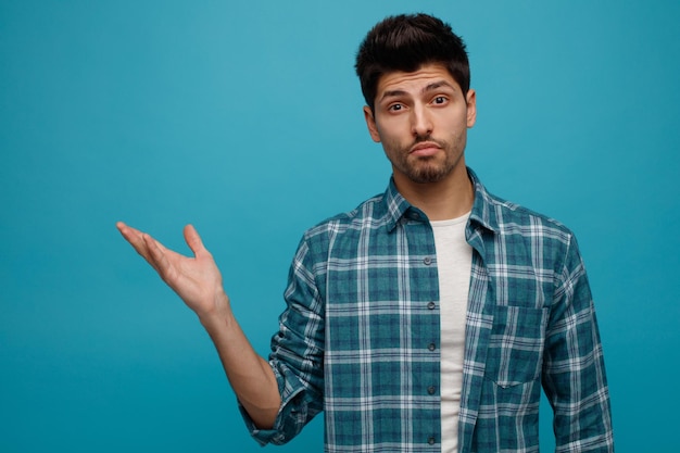 Unsure young man looking at camera showing empty hand isolated on blue background