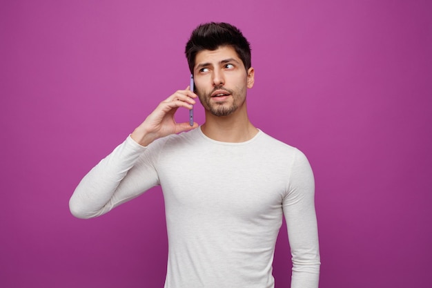 Unsure young man looking at side talking on phone isolated on purple background