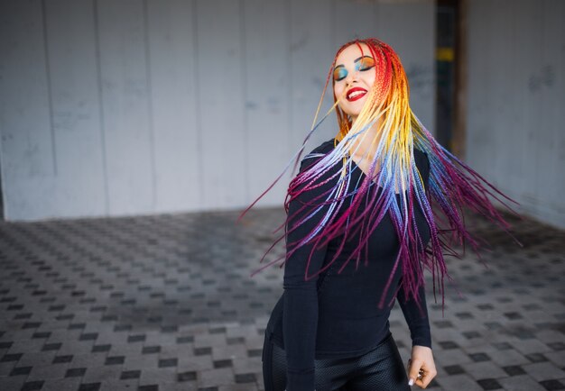 Unusual elegant girl in a black simple dress with bright makeup and multi-colored braids posing against the background of an old building