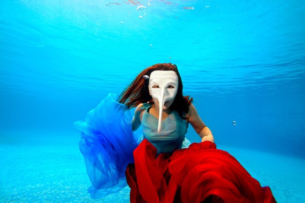 An unusual girl underwater in a white carnival mask poses on a blue background with a red cloth