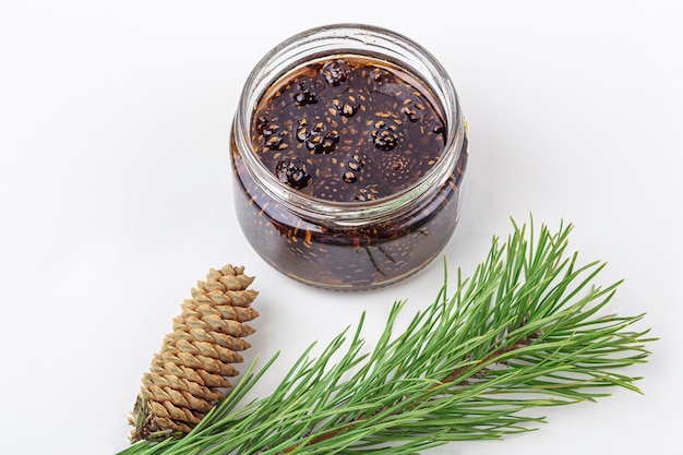 Unusual jam from pine cones in glass jar on white background among pine branches and cones. Organic and vegetarian sweet dessert close up, copy space for text. Remedy for colds