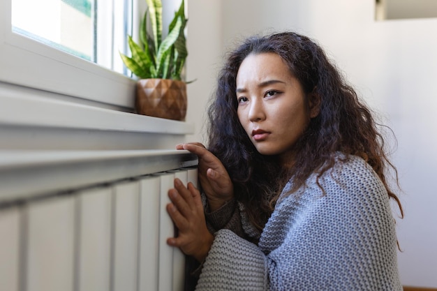 몸이 좋지 않은 아시아 여성 임차인은 담요를 덮고 있는 차가운 거실에 앉아 오래된 라디에이터에 손을 얹고 열 부족으로 고통받고 있습니다.