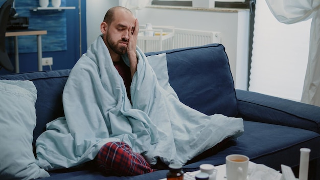 写真 家で毛布に包まれた頭痛を持っている体調不良の男。風邪やインフルエンザの症状がこめかみをこすり、薬や薬瓶をテーブルに置いて病気を治そうとしている病気の人。
