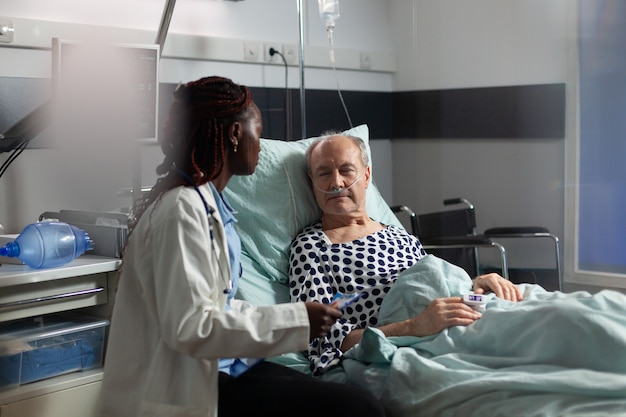 Unwell senior patient laying in bed breathing through oxygen test tube, listening to doctor