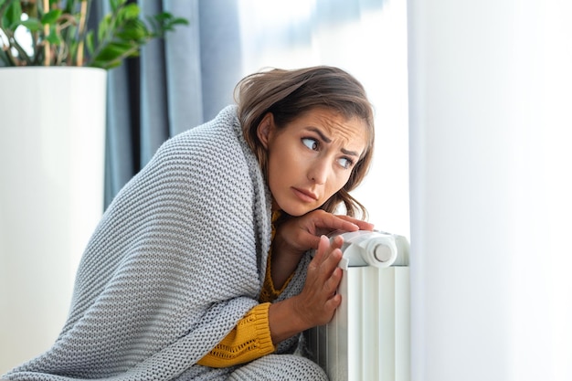 Unwell woman renter in blanket sit in cold living room hand on old radiatorsuffer from lack of heat Unhealthy young woman struggle from chill freeze at home No heating concept