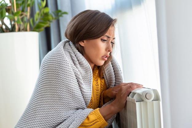 Unwell woman renter in blanket sit in cold living room hand on old radiatorsuffer from lack of heat Unhealthy young woman struggle from chill freeze at home No heating concept