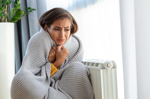 Unwell woman renter in blanket sit in cold living room hand on old radiatorsuffer from lack of heat Unhealthy young woman struggle from chill freeze at home No heating concept