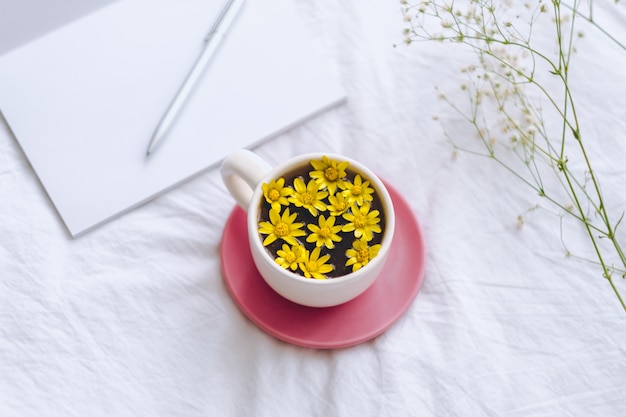 Ð¡up with yellow flowers inside, on a white bed with notepad and pen.