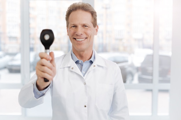 Upbeat experienced friendly doctor enjoying working hours in the clinic and showing dermatoscope while expressing positivity