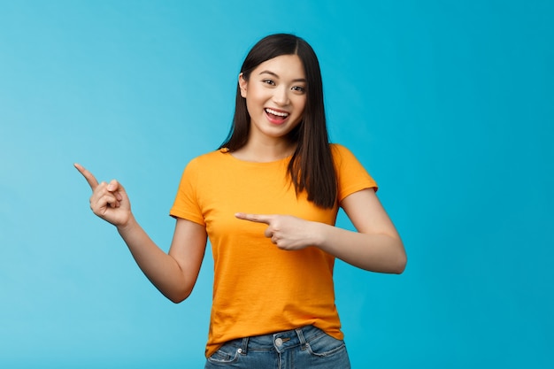 Upbeat friendly asian cute girl with dark haircut lively pointing left introduce awesome new copy space, smiling broadly toothy happy grin, stand blue background joyful, recommend link.