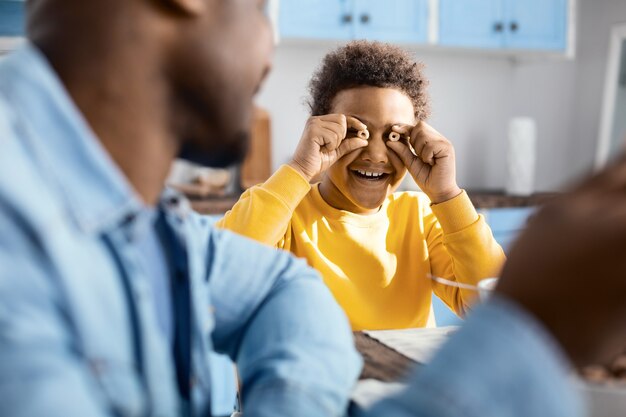 Upbeat mood. The focus being on a cheerful curly-haired boy goofing around during breakfast and smiling happily while holding two cereal rings near his eyes