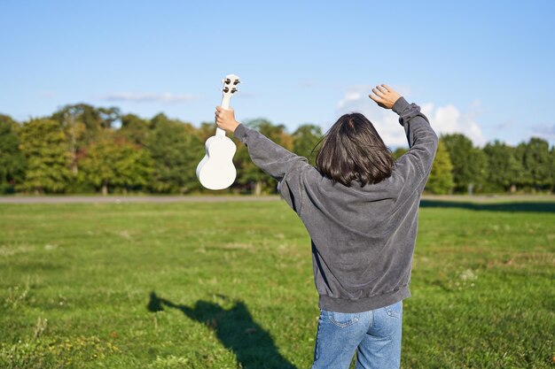 Photo upbeat young woman dancing with her musical instrument girl raises her ukulele up and pose in park o