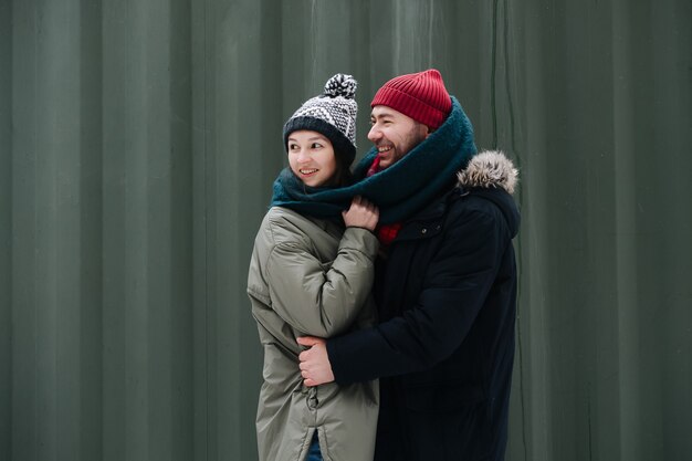 Uplifted couple in love standing outdoors at winter, looking at someone. Both wearing hats, jackets and scarfs. In front of corrugated sheet house wall.