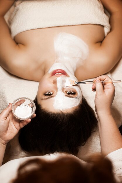 Upper view of a charming woman leaning on a bed looking at the camera while doing a white mask on her face and neck in a wellness center.